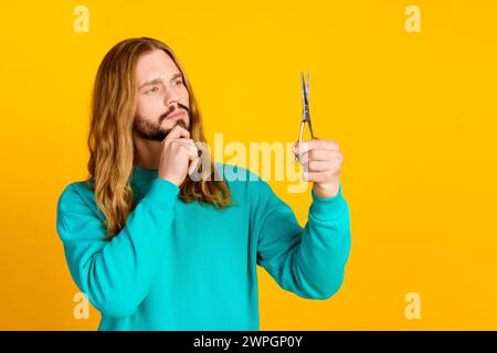 Foto des nachdenklichen Mannes Armberührung Kinnblick Schere halten Brainstorming leeren Raum isoliert auf gelbem Hintergrund Stockfoto