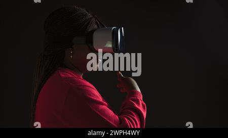 Seitenansicht einer kreativen jungen Frau, die mit interaktiver Technologie mit einer 3D-Virtual-Reality-Brille simuliert wird. Afrikanisch-amerikanische Fashionista mit einem hochentwickelten VR-Headset. Stockfoto
