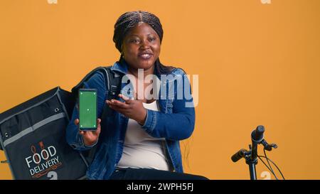 Weibliche afroamerikanische Kurierin, die ein digitales Gerät mit grünem Bildschirm hält, während sie auf dem Fahrrad sitzt. Junge Frau, die ein Smartphone mit leerem Modell-Copyspace-Template zeigt. Stockfoto