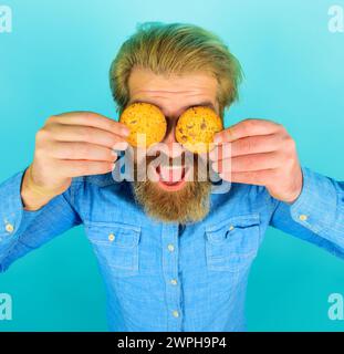 Bärtiger Mann bedeckte die Augen mit frisch gebackenen Haferflockenkeksen. Leckerer süßer Snack. Gutaussehender Mann isst Schokoladenkekse Kekse. Frühstück oder Mittagessen Stockfoto