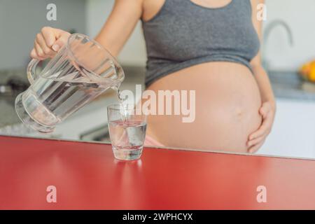 Eine schwangere Frau, die die lebenswichtigen Vorteile des Wassers während der Schwangerschaft berücksichtigt, steht mit einem Glas in der Küche und unterstreicht die entscheidende Rolle der Hydratation bei Stockfoto