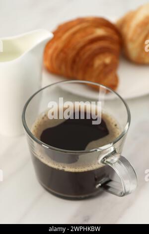 Aromatischer Kaffee in Glasbecher, Krug und frische Croissants auf weißem Marmortisch, Nahaufnahme Stockfoto