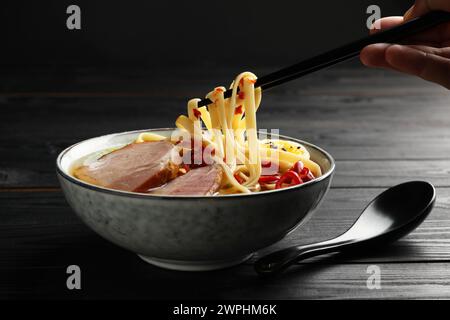 Frau isst köstliche Ramen mit Stäbchen an schwarzem Holztisch, Nahaufnahme. Nudelsuppe Stockfoto