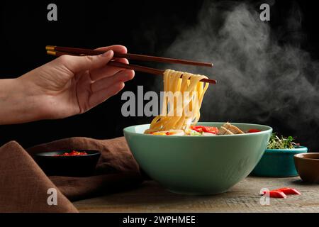 Frau isst köstliche Ramen mit Stäbchen am Holztisch, Nahaufnahme. Nudelsuppe Stockfoto