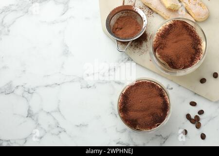 Köstliches Tiramisu in Gläsern, Keksen, Kaffeebohnen und Sieb mit Kakaopulver auf weißem Marmortisch, Blick von oben. Leerzeichen für Text Stockfoto