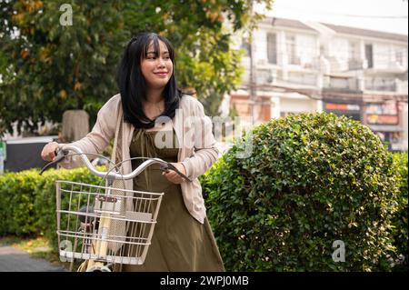 Eine schöne, positive junge asiatische Frau in einem süßen Kleid, die wegschaut und mit ihrem Fahrrad auf dem Fußweg in der Stadt spaziert. Stockfoto