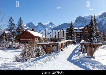 Holzfußgängerbrücke Spring Creek Mountain Village, Canmore, Alberta. Die Skyline Von Three Sisters Canadian Rocky Mountain Peaks Wurde Von Fernschnee Bedeckt Stockfoto