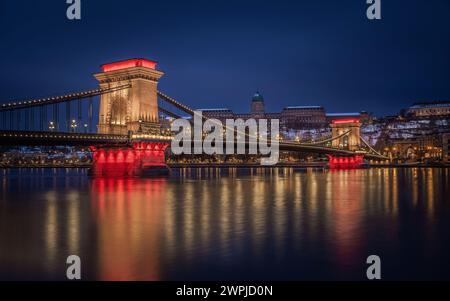 Budapest, Ungarn - beleuchtete Szechenyi Kettenbrücke in der Abenddämmerung an einem Winterabend mit dem Königspalast von Buda im Hintergrund mit den schneebedeckten Budaer Hügeln A Stockfoto