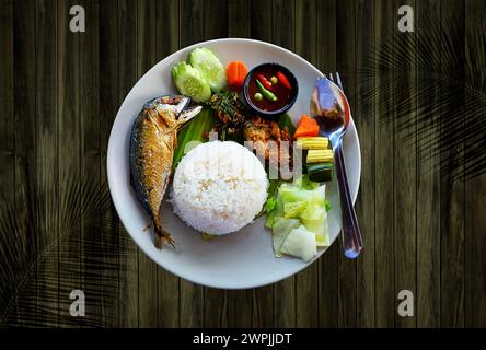 Thailändischer Reis, Chilipaste, Garnelenpaste, gebratene Makrele, Gemüse auf einer weißen Keramikplatte auf hölzernem Hintergrund. Stockfoto