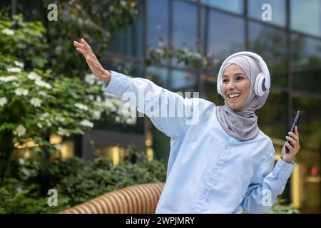 Junge muslimische Studentin außerhalb des Universitätsgeländes, fröhlich tanzend vor dem Gebäude, Frau im Hijab, die Musik mit Telefon und Kopfhörern hört, h Stockfoto