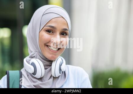 Porträt einer lächelnden jungen muslimischen Frau, die einen Hijab und Kopfhörer um den Hals trägt und das Glück in einer städtischen Umgebung zum Ausdruck bringt. Stockfoto