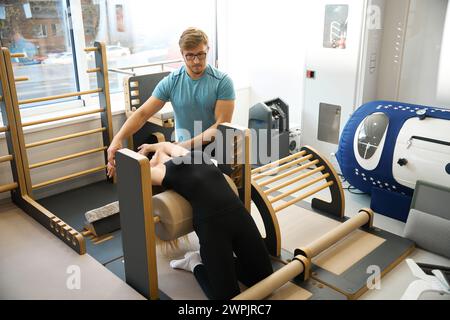 Mitarbeiter der Rehabilitationsklinik hilft Frauen bei der Arbeit an einem speziellen Simulator Stockfoto