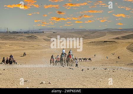 Gizeh Nekropolis, Ägypten - 26. April 2022: Touristen reiten in der Wüste auf Kamelen. Im Hintergrund der laufende Bau des zukünftigen größten Histos Stockfoto