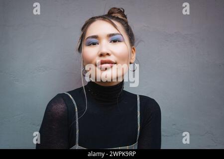 Asiatische Frau im urbanen Stil posiert selbstbewusst - auffälliges Make-up und minimalistische Mode vor grauem Hintergrund Stockfoto
