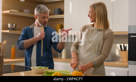 Kaukasische Frau schneidet Gemüse kochen Salat Mann mittleren Alters Ehemann geben Löffel zu Frau erwachsenes Paar glückliche Familie singen Lied und Spaß beim Narren Stockfoto