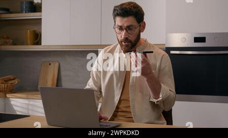 Multitasking Kaukasischer Mann, der mit Laptop-Aufnahme arbeitet Diktieren von Sprachnachrichten Voicemail mit Telefon in der Küche zu Hause. Männlicher freiberuflicher Geschäftsmann Stockfoto