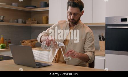 Kaukasischer Mann freischaffender Geschäftsmann Benutzer mit Laptop arbeiten von zu Hause entfernt Internet Computer Arbeit in der Küche gießen Wasser von der Flasche ins Glas Stockfoto