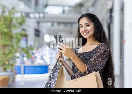 Fröhliche indische Käuferin, die ihr Handy benutzt, während sie bunte Taschen in einem modernen Einkaufszentrum hält. Stockfoto