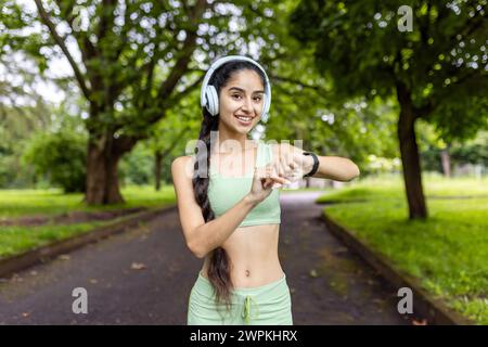 Lächelnde junge Indianerin in Sportbekleidung mit Kopfhörern, die ihr Workout in einer üppigen Parklandschaft mit Smartwatch überwacht. Stockfoto