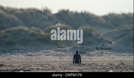 Sonnenlicht am frühen Morgen über Antony Gormleys „Another Place“ am Crosby Beach mit einer grasbewachsenen Sanddüne als Hintergrund Stockfoto