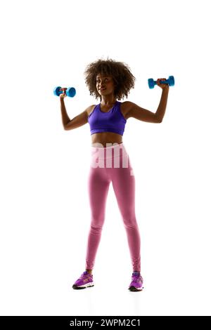 Lächelnde Frau mit hochgezogenen Kurzhanteln, die einen violetten Sport-BH und Leggings vor weißem Studiohintergrund trägt. Stockfoto