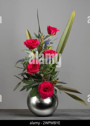 Rote Rosen, angeordnet in einer kugelförmigen Spiegelvase, vor grauem Hintergrund Stockfoto
