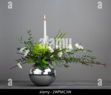 Blumenanordnung einer Kerze in einem kugelförmigen Spiegelkugelbehälter auf grauem Hintergrund. Stockfoto