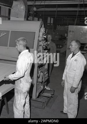 1950er Jahre, historisch, zwei männliche Arbeiter in Overalls in einer Produktionshalle in der Papierfabrik Camas, einer leitete einen jüngeren Arbeitskollegen, während er Holzbretter in ein Maschinenstück bei der Crown Zellerbach Corporation, WA, USA, einspeist. Stockfoto