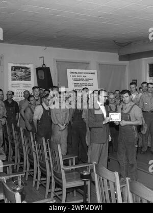 1950er Jahre, historisch, in einer Halle versammelte sich eine Gruppe amerikanischer Papierfabrikarbeiter, um zu beobachten, wie ein männlicher Arbeiter in dämlichen Dunjarees einen Gesundheits- und Sicherheitspreis von einem Firmenleiter mit Fliege, WA, USA, erhält. Ein Schild an der hinteren Wand besagt... Camas Mill Unfallverhütungsrichtlinien. 1. Grundlegender Sicherheitsplan; 1) Endziel ist die vollständige Beseitigung aller Verletzungen...... Stockfoto