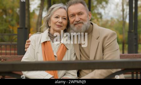 Romantisches Paar kaukasische reife Frau Mann liebt es, sich draußen im Park zu entspannen, lächelnd, friedlich, alte Ehegatten, Ehemann, der sich auf der Bank umarmt Stockfoto