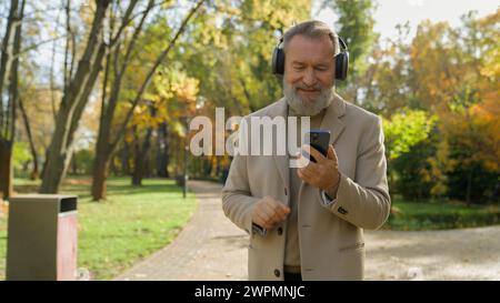 Senior Kaukasier Mann 60er Jahre alt männlich spazieren im Herbst Park Stadt Musik hören in kabellosen Kopfhörern tanzen Musikvideo auf Handy glücklich Stockfoto