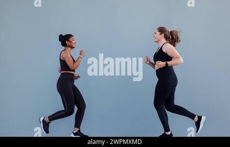 Sportlerin, die vor ihrem Freund läuft und zum Laufen motiviert. Zwei Joggingfreunde lächeln, während sie an der grauen Wand rennen. Sportlerin, die vorne läuft Stockfoto