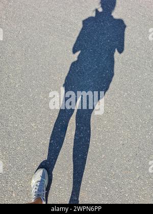 Nur der Schatten einer Frau und ihr linker weißer Sportschuh sind auf der Straßenoberfläche zu sehen Stockfoto