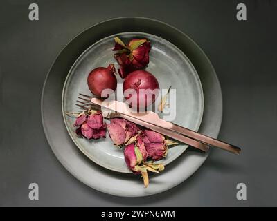 Anordnung von zwei ganzen roten Zwiebeln mit getrockneten Rosenblättern neben kupferfarbenem Besteck auf einer Porzellanplatte als Stillleben Stockfoto