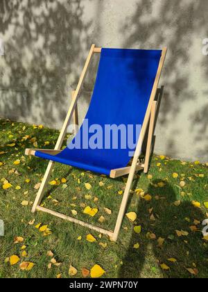 Ein blauer Liegestuhl steht im Sonnenlicht auf einem grünen Rasen vor einer hellgrauen Betonwand mit Schatten von Blättern Stockfoto