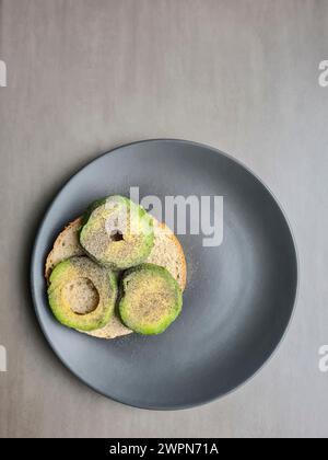 Eine in Scheiben geschnittene Avocado, die mit Pfeffer gewürzt wurde, liegt auf einem halben Brötchen auf einem grauen Teller vor grauem Hintergrund Stockfoto