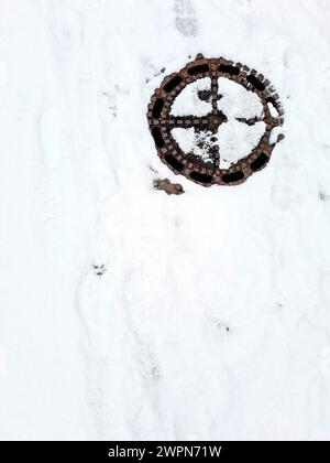 Spuren und Muster im Schnee und die schneebedeckte Mannlochabdeckung schaffen ein abstraktes Winterbild Stockfoto