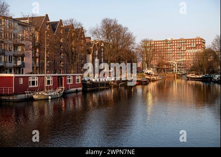 Amsterdam Niederlande 8. März 2024 Westelijke Eilanden. Bei Sonnenuntergang spiegeln sich die Gebäude des Realeneilands im glitzernden Wasser des Kanals. centrum, pakhuizen, Pakhuis, Dämmerung, Avondschemer, gracht, Grachtenpand, Hausboote, Woonboten, Woonark, Stockfoto