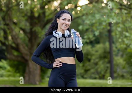 Eine begeisterte Fitnessbegeisterte erholt sich in einem grünen Park und hält eine Wasserflasche mit Kopfhörern um den Hals. Stockfoto