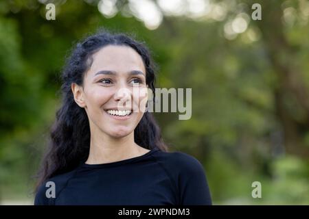 Porträt einer zufriedenen jungen Frau, die sanft lächelt, mit grünen Bäumen im Hintergrund, die Glück und Ruhe darstellen. Stockfoto
