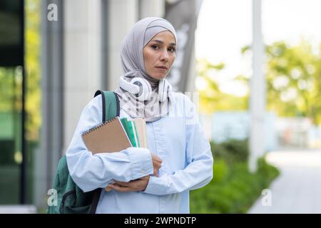 Porträt einer fokussierten muslimischen Frau mit Hijab, Kopfhörern und Lehrbüchern, die in einem akademischen Umfeld nach draußen treten. Stockfoto
