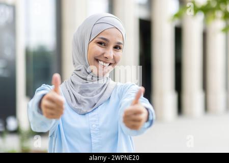 Eine fröhliche junge Frau, die einen Hijab trägt, zeigt mit einem breiten, selbstbewussten Lächeln zwei Daumen hoch und signalisiert Anerkennung und Erfolg. Stockfoto