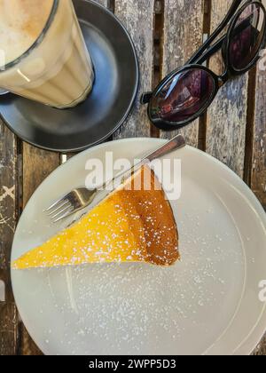 Ein Stück Käsekuchen mit Puderzucker auf einer weißen Platte Kaffee mit Milch im Glas zum Nachtisch, schwarze Sonnenbrille auf einem Holztisch Stockfoto