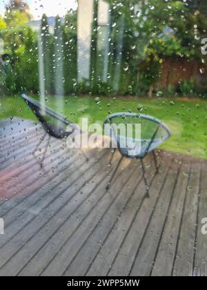 Blick durch die Fensterscheibe mit Regentropfen auf die nasse Terrasse mit zwei modernen Gartenstühlen Stockfoto