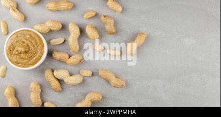 Draufsicht. Verstreute rohe Erdnüsse und ein Glas Erdnussbutter auf grauem Hintergrund. Stockfoto