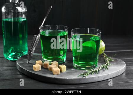 Absinth in Gläsern, Rosmarin, brauner Zucker und Limette auf schwarzem Holztisch. Alkoholisches Getränk Stockfoto