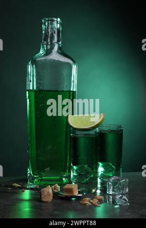 Absinth in Shot Gläser mit Eiswürfeln, Limette, braunem Zucker und Löffel auf grauem Tisch vor grünem Hintergrund. Alkoholisches Getränk Stockfoto