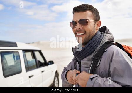 Mann, Lächeln und Rucksack in der Sanddüne für Wanderungen, Entdeckungen oder Outdoor-Abenteuer in der Natur. Mann, Sonnenbrille und Glück mit Campingausrüstung für Stockfoto