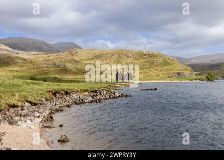 Das ruinierte Calda House aus dem achtzehnten Jahrhundert steht jenseits eines felsigen Ufers von Loch Assynt, Schottland, mit grasbewachsenen Hügeln unter grauen Wolken im Hintergrund. Stockfoto