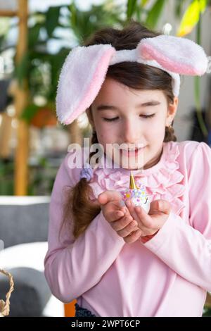 Ein süßes Mädchen mit rosa Hasenohren macht ein Osterbasteln - dekoriert ein Ei in Form eines Einhorns mit Strasssteinchen, Horn, Blumen im Inneren Stockfoto